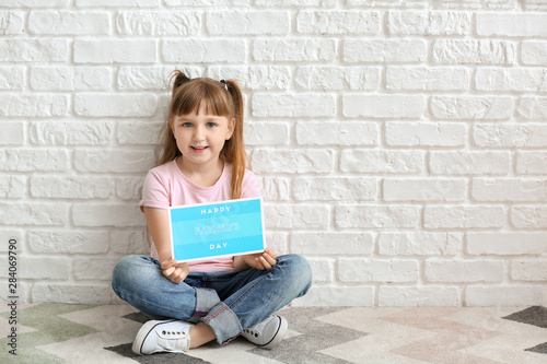 Cute little girl with greeting card for Father's Day sitting near white brick wall