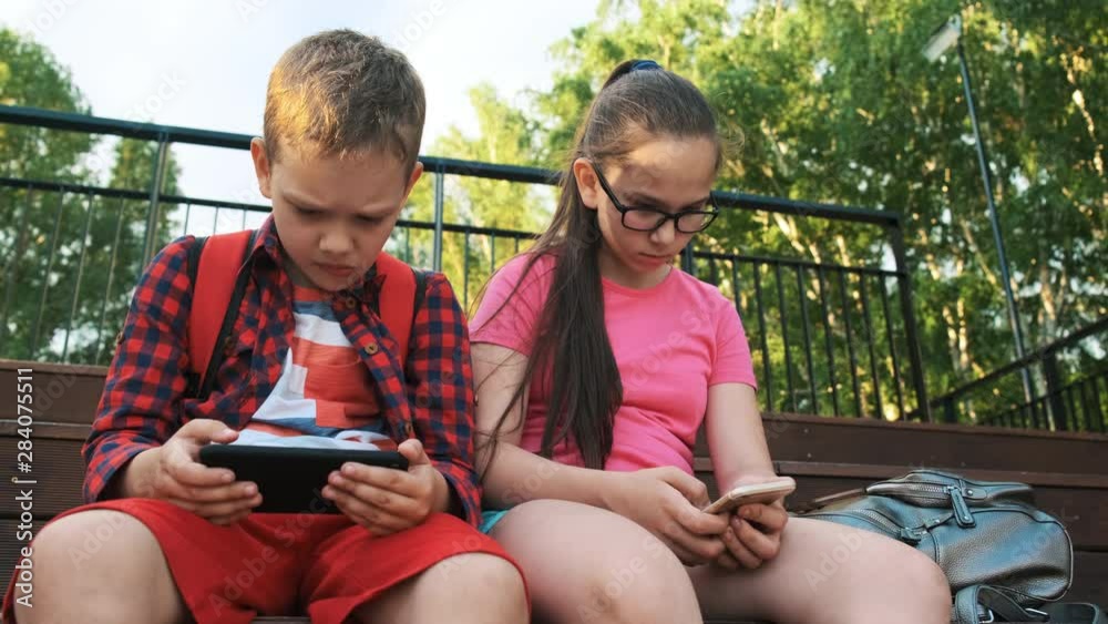 The older sister and brother are sitting on the street, looking at smartphones. A boy plays in a mobile application, a girl looks at a photo. Children walk after school The older sister and brother are sitting on the street, looking at smartphones. A boy plays in a mobile application, a girl looks at a photo. Children walk after school