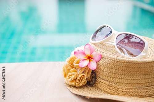 Summer concept background, design sunglasses on brown hat with beautiful flower over blurred blue water background, outdoor day light