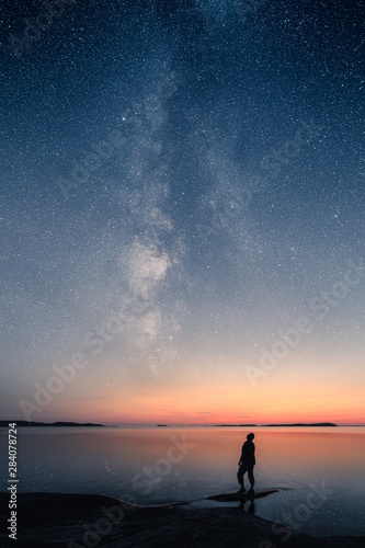 Фотография A man standing by a calm sea and looking up on the stars of the milky way