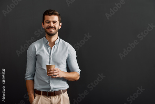 Drinking cappuccino. Cheerful young bearded man holding a cup of hot coffee and looking at camera with smile while standing against dark background photo