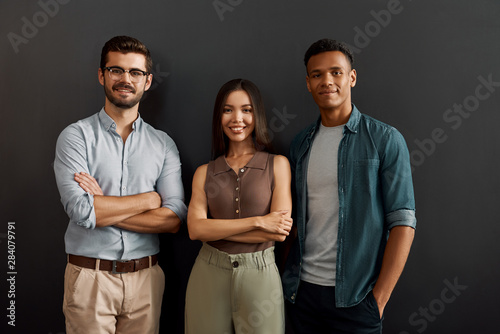 We are great team. Group of three multicultural cheerful young people in casual wear looking at camera with smile while standing against dark background photo