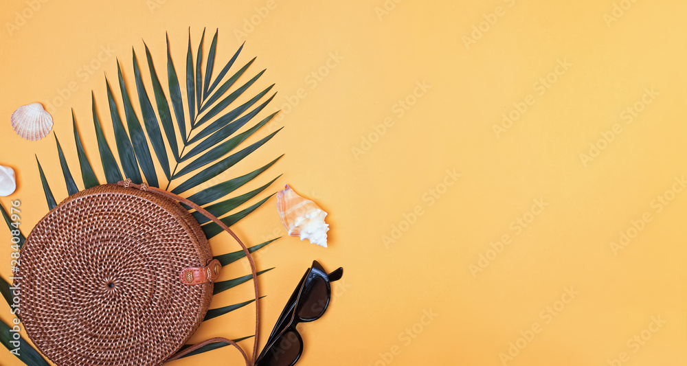 Fototapeta premium Stylish round straw handbag with palm leaf, sunglasses and seahells on the bright yellow background,