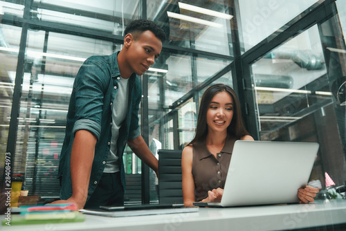 Multiracial team. Young asian woman and afro american man in casual wear looking at screen of laptop and discussing new project while working in modern office