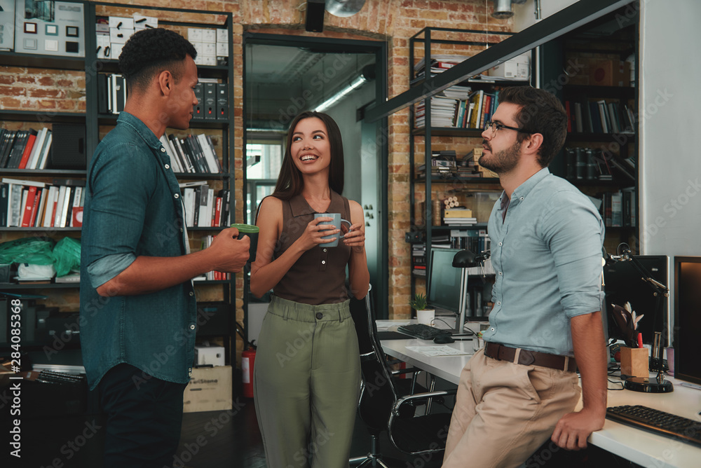 Nice coffee! Young smiling colleagues in casual wear holding cups and talking about something while standing in modern office