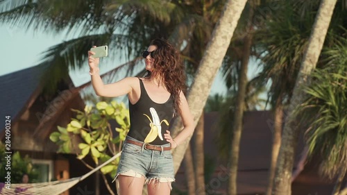 Pretty young woman in sunglasses and swimsuit takes selfie on beautiful sandy beach by sea. Happy smiling girl pulls her hair, spinning and taking pictures of herself with smartphone. Slow motion.