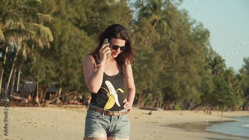 Pretty young woman in sunglasses and swimsuit makes phone call and talks walking on sandy beach. Happy girl on vacation walks along sea shore of tropical island and calls on smartphone on sunset.