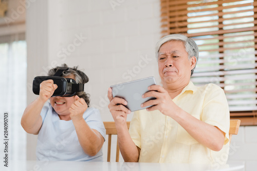 Asian elderly couple using tablet and virtual reality simulator playing games in living room, couple feeling happy using time together lying on table at home. Lifestyle Senior family at home concept.