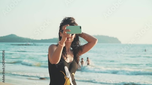 Pretty young woman in sunglasses and swimsuit takes selfie on beautiful sandy beach by sea. Happy smiling girl pulls her hair, spinning and taking pictures of herself with smartphone. Slow motion.