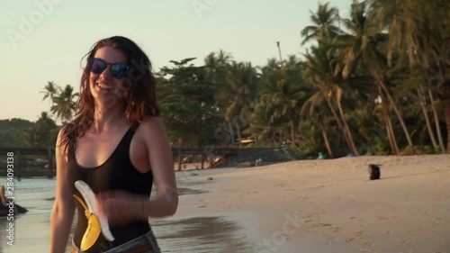 Young happy caucasian woman is spinning and dancing on beach of tropical island at sunset. Smiling pretty girl in sunglasses enjoys freedom and vacation on sandy coast by sea. Slow motion footage.