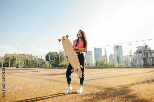 Beautiful curly . girl in casual clothes stands with a longboard in hands on a tennis court, looks in camera and poses.Hispanic girl with skateboard in hands posing on playground.Street culture.
