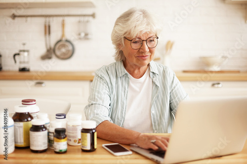 Positive beautiful gray haired female pensioner in spectacles choosing healthy lifestyle, sitting in kitchen with dietary supplements, keyboarding on laptop, typing review via online store