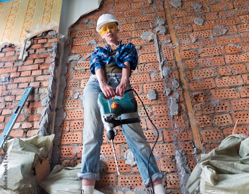 young woman in work clothes and a protective helmet stands next to a brick wall and holds a heavy hammer drill