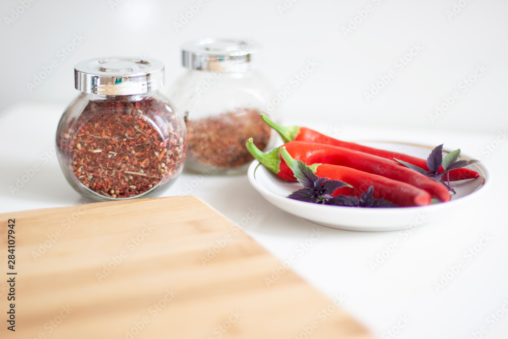 Red hot peppers on a white plate with basil leaves. Cutting kitchen board in the foreground and jars of spices in the background Red hot peppers on a white plate with basil leaves. Cutting kitchen board in the foreground and jars of spices in the background