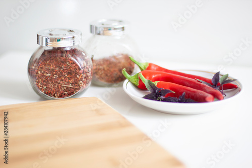 Red hot peppers on a white plate with basil leaves. Cutting kitchen board in the foreground and jars of spices in the background