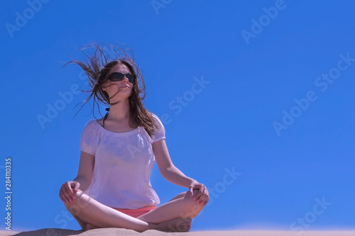 Woman in sunglasses is meditating on vacation. Travel tourist sitting at seashore. Girl meditate looks around sandy beach on blue sky background yoga lotus pose. Traveller tourism in summer wind blow.