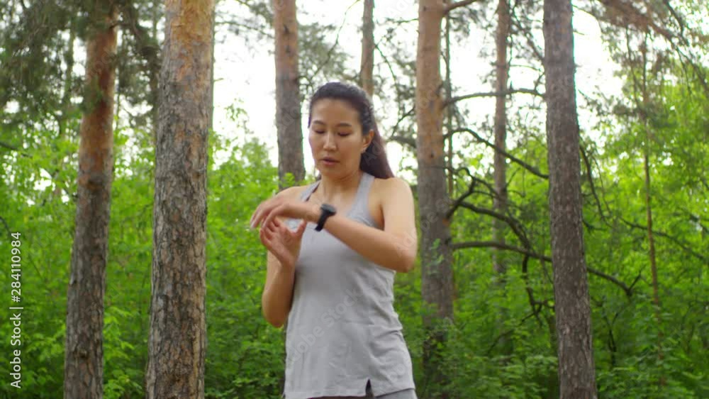 Lockdown shot of young Asian woman with long black hair in ponytail, wearing grey vest and shorts, running through forest, stopping, looking at watch, wiping sweat off forehead, and jogging away Lockdown shot of young Asian woman with long black hair in ponytail, wearing grey vest and shorts, running through forest, stopping, looking at watch, wiping sweat off forehead, and jogging away