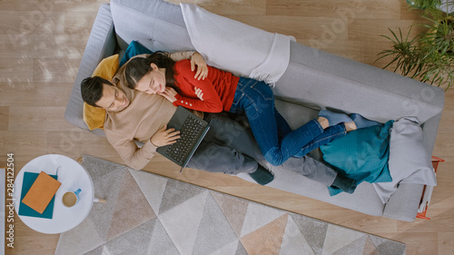 Young Couple is Lying Down on a Couch, Using a Laptop. They are Happy and Smile. Cozy Living Room with Modern Interior with Plants, Table and Wooden Floor. Top View.