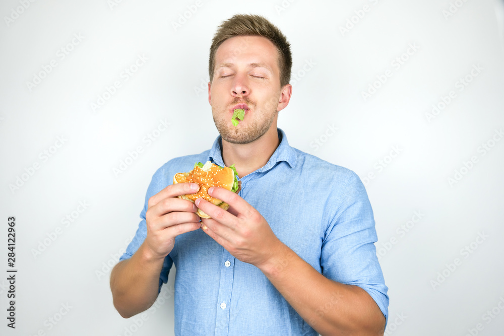 young funny man biting cheeseburger from fast food restaurant standing with salad leaf in his mouth on isolated white background