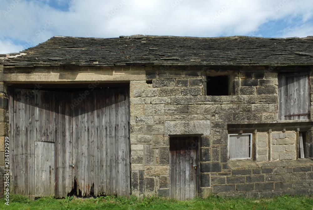 ancient abandoned stone barn in a row of rural buildings with empty windows and wooden doors with the pavement overgrown with grass ancient abandoned stone barn in a row of rural buildings with empty windows and wooden doors with the pavement overgrown with grass