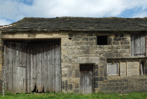 ancient abandoned stone barn in a row of rural buildings with empty windows and wooden doors with the pavement overgrown with grass