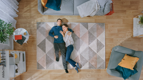 Young Happy Couple is Lying Down on the Floor and Laughing. Man Hugs the Girl. Cozy Living Room with Modern Interior with Carpet, Sofa, Chair, Table, Shelf, Plants and Wooden Floor. Top Down.