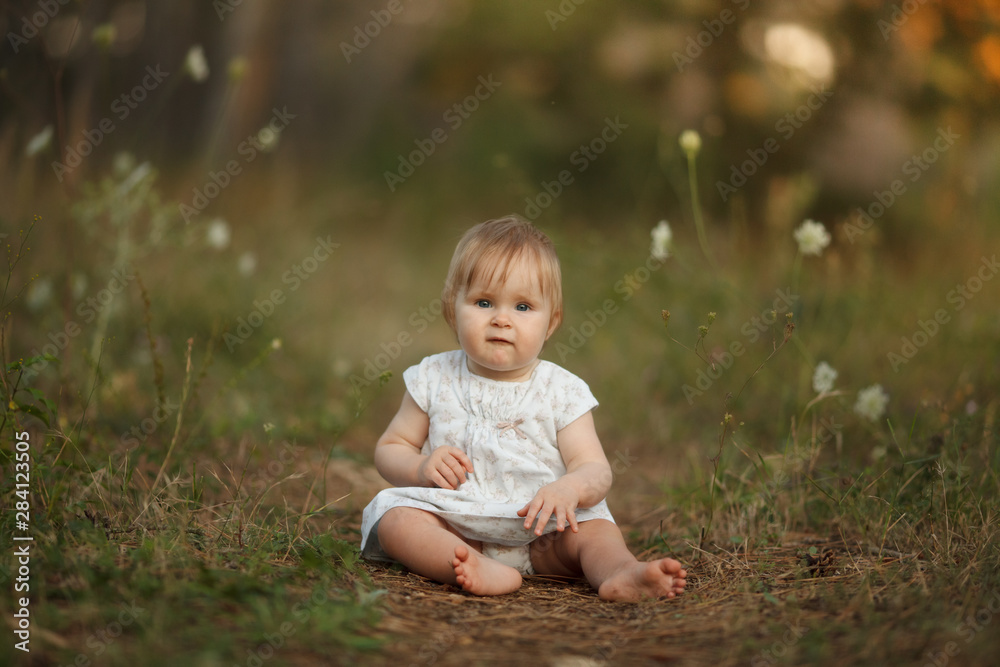 A cute little baby girl in a light dress is sitting on a path in the forest. Looking into the camera. The concept is tenderness, naivety, simplicity, family, motherhood. A cute little baby girl in a light dress is sitting on a path in the forest. Looking into the camera. The concept is tenderness, naivety, simplicity, family, motherhood.