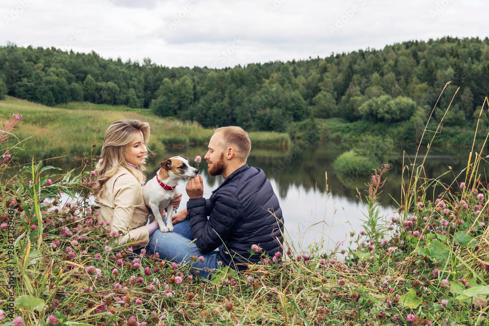 a young couple sitting on the shore of the lake and playing with a dog breed Jack Russell Terrier, they are smiling and happy