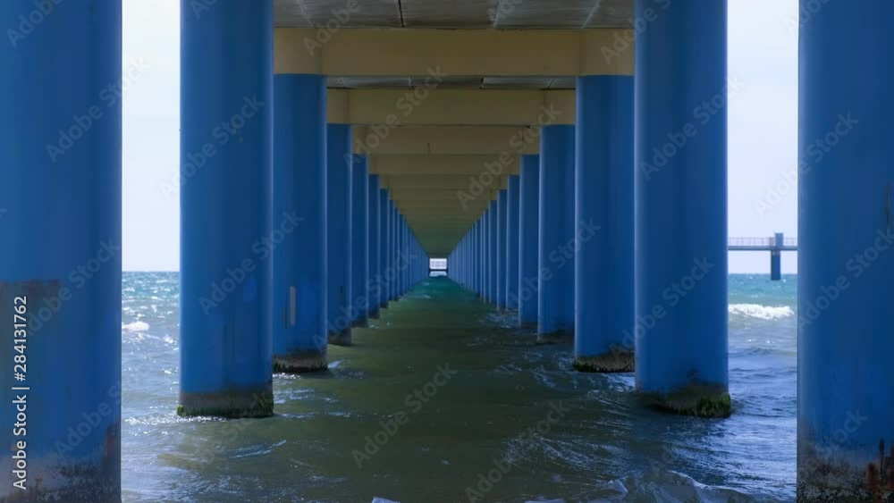 Sea under pier among parallel blue columns making narrow corridor in water. Seascape through pier with sea waves and blue sky at sunny day. Sea under pier among parallel blue columns making narrow corridor in water. Seascape through pier with sea waves and blue sky at sunny day.