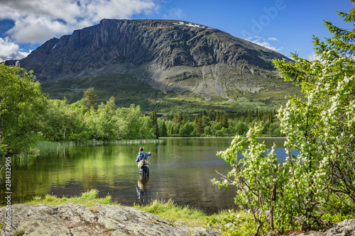 Angler beim Fliegenfischen mit Angelroute im Wasser stehend mit Wathose im See vor Bäumen und Berg