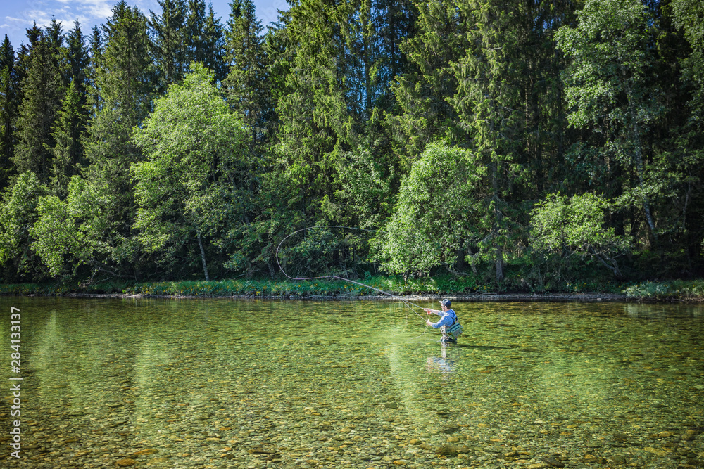 Angler beim Fliegenfischen mit Angelroute im Wasser stehend mit Wathose im Fluss vor Bäumen