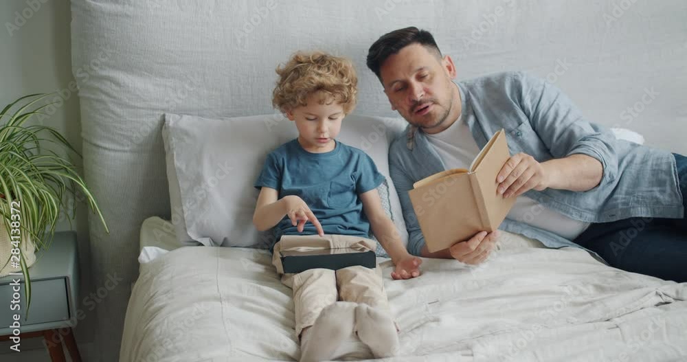 Loving father middle-aged man is reading book to cute child while kid is using tablet touching screen in bed in apartment. Family, literature and gadgets concept. Loving father middle-aged man is reading book to cute child while kid is using tablet touching screen in bed in apartment. Family, literature and gadgets concept.