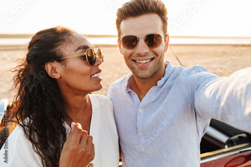 Photo of fashionable multiethnic couple wearing sunglasses taking selfie photo together while standing by car on beach © Drobot Dean