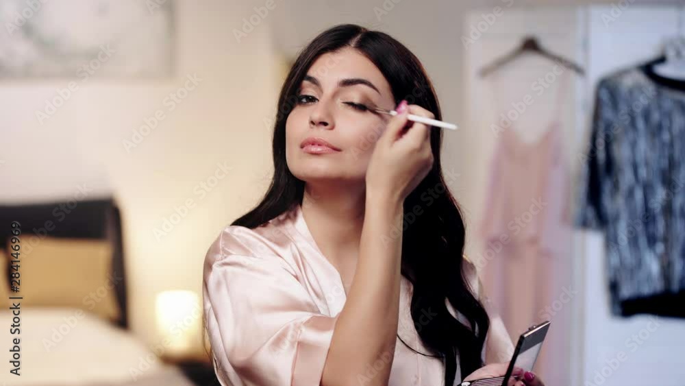 brunette woman in satin robe applying eye shadow, evening makeup
