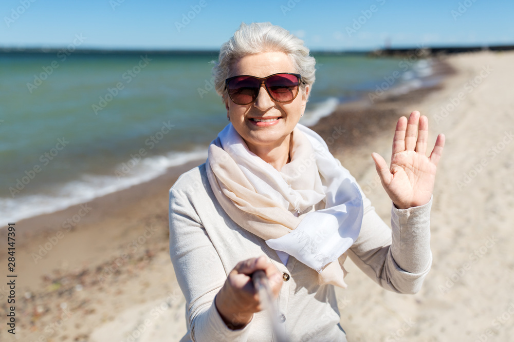 old people and leisure concept - happy smiling senior woman in sunglasses taking picture by selfie stick and waving hand on beach in estonia
