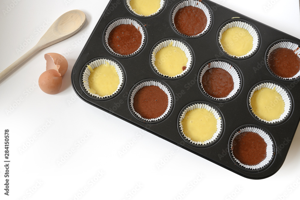 Naklejka premium muffin baking tray filled with light and dark dough for delicious cupcakes, background fades to white, copy space, high angle view from above
