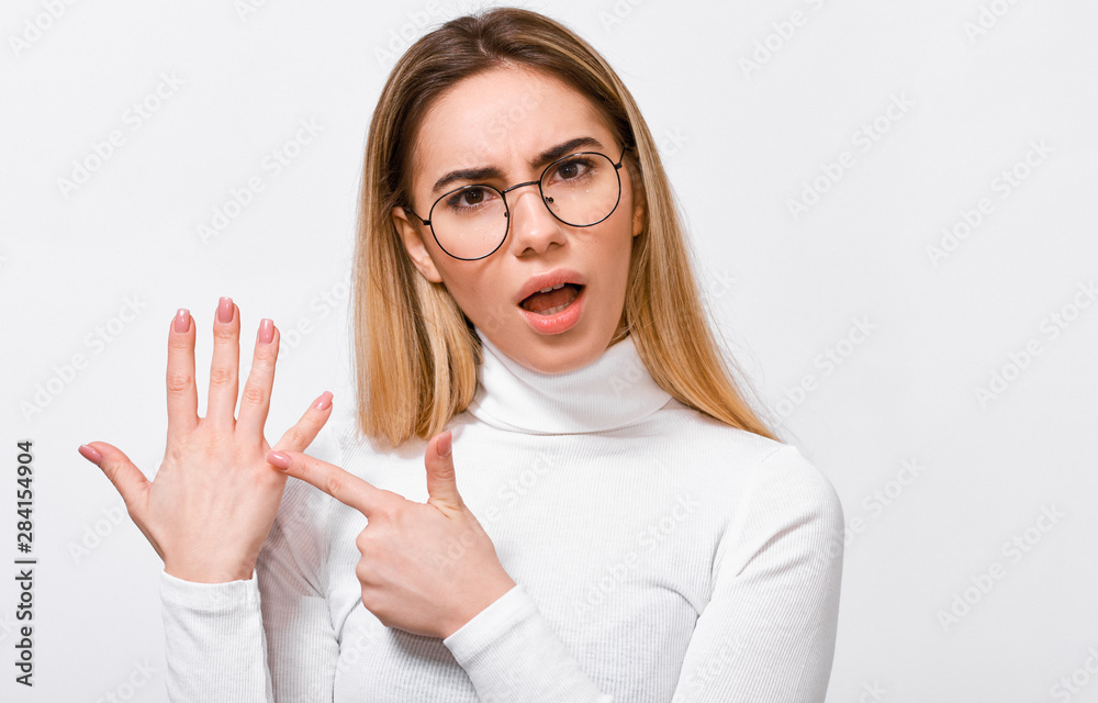 Upset young blonde woman wearing white blouse and round trendy transparent glasses, showing engagement ring on her finger, posing over white studio background. Upset young blonde woman wearing white blouse and round trendy transparent glasses, showing engagement ring on her finger, posing over white studio background.