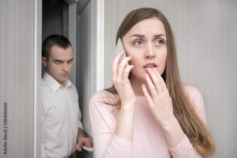 Frightened young woman calls the rescue service, her husband enters the room and looks at her angrily, relationships concept Frightened young woman calls the rescue service, her husband enters the room and looks at her angrily, relationships concept
