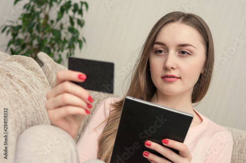 Attractive woman uses tablet, holding a plastic card, sitting at home on the couch, toned