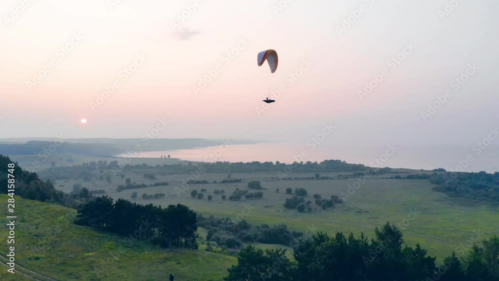 A person on a parawing is gliding above the green landscape A person on a parawing is gliding above the green landscape