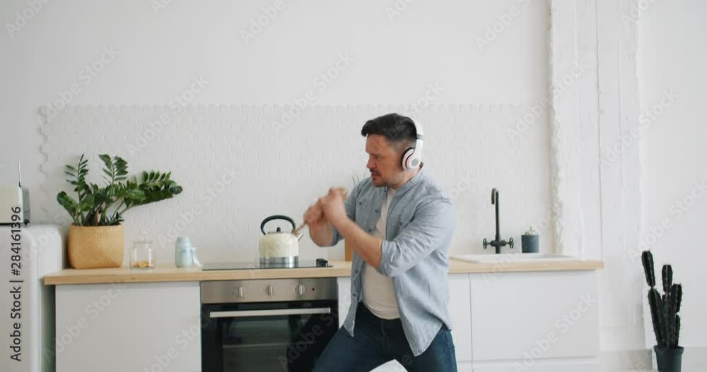 Silly man in headphones is singing in spoon in kitchen dancing playing imaginary guitar having fun alone in apartment. People and modern lifestyle concept. Silly man in headphones is singing in spoon in kitchen dancing playing imaginary guitar having fun alone in apartment. People and modern lifestyle concept.