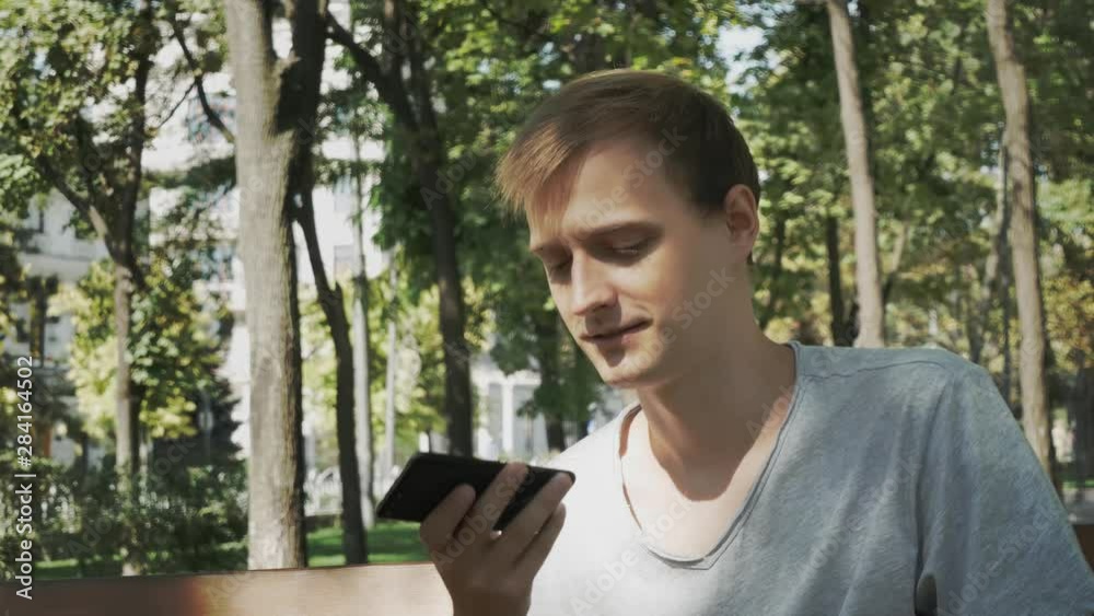 Happy man using a smart phone voice recognition audio ai message speech function on line walking on a street with city background, speech message helper student male in park on bench Happy man using a smart phone voice recognition audio ai message speech function on line walking on a street with city background, speech message helper student male in park on bench