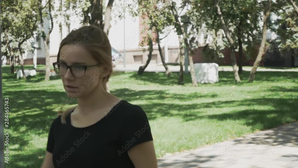 Pretty young caucasian woman in eyeglasses walking towards camera, checking phone and celebrating good news in a park Pretty young caucasian woman in eyeglasses walking towards camera, checking phone and celebrating good news in a park