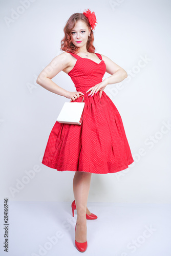 Beautiful redheaded girl in red midi pinup style dress isolated on white studio background standing alone