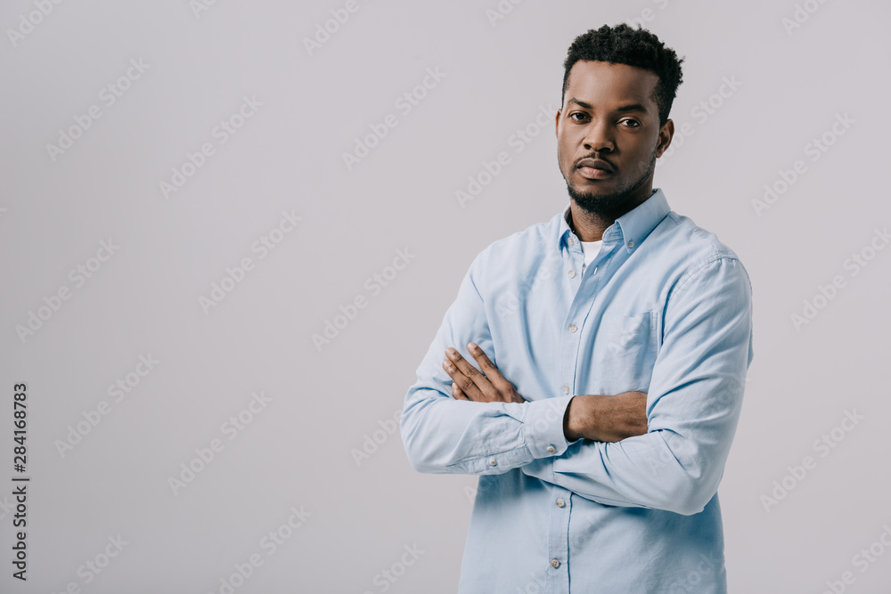 handsome african american man looking at camera and standing with crossed arms isolated on grey