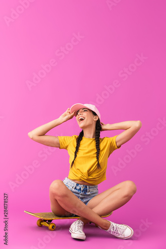 Image of happy caucasian woman wearing cap laughing and sitting on skateboard © Drobot Dean