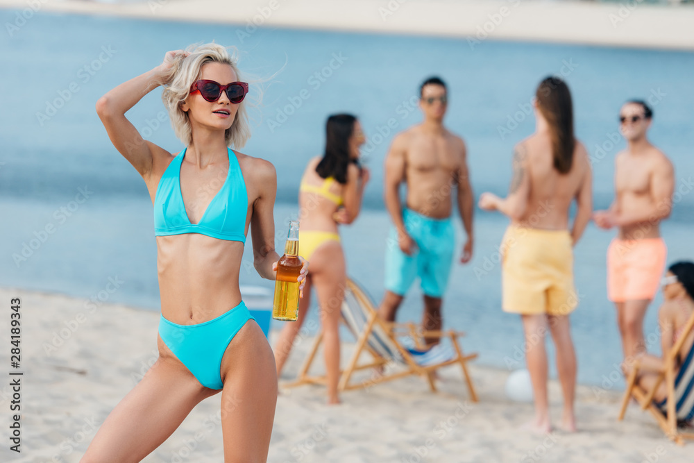beautiful young woman in swimsuit holding bottle of beer and looking away while standing on beach near multicultural friends