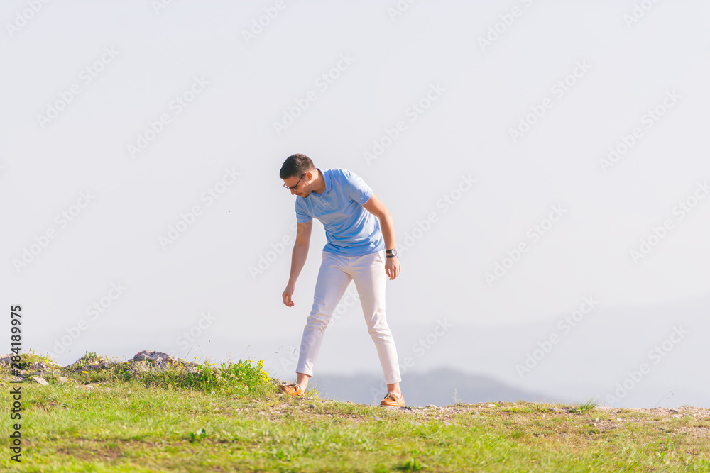Well dressed ( fashionable) man stands in nature looking over a cliff at the large lake and mountain line while wearing boat shoes, polo shirt and formal pants.