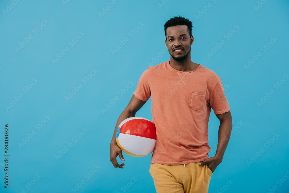 happy african american man standing with hand in pocket and holding beach ball isolated on blue