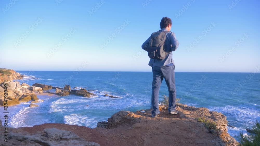 A backpacker man watching the ocean from the rock of a cliff clif A backpacker man watching the ocean from the rock of a cliff clif